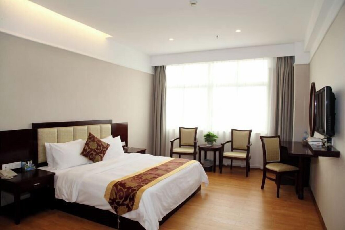 A hotel room with a large bed, two chairs, a small table, a TV, and a window with curtains.