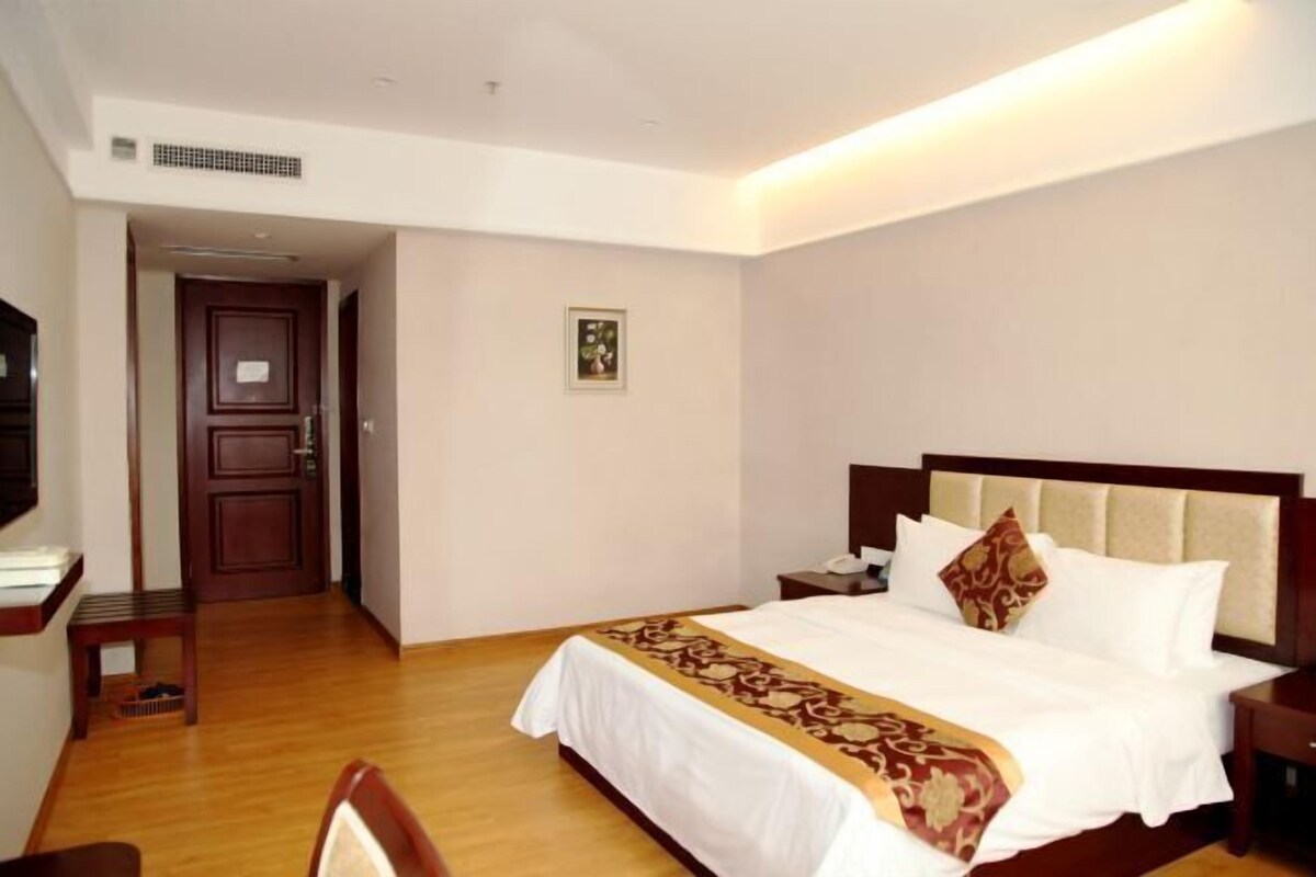 A hotel room with a wooden floor, a bed with white bedding and a brown decorative throw, a nightstand with a lamp, and a wooden door.