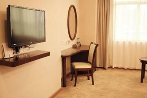 A hotel room with a flat-screen TV mounted on a wall, a desk with a chair, a round mirror, and a window with curtains. - Jinjue Hotel (Nearby Train Station) (Huizhou)
