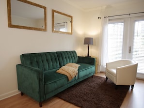 Un sofá de terciopelo verde con una manta, un sillón blanco, una lámpara y dos espejos en la pared.