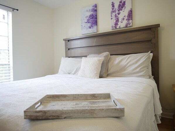 Un lit bien fait, avec un plateau en bois placé devant, une tête de lit et deux tableaux floraux accrochés au mur.