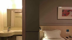 A hotel room with a bed, a bedside table with a phone, a wall-mounted painting, and a bathroom with a sink and mirror. - Jincheng Hotel (Bazhou Shengfang Town) (Bazhou)
