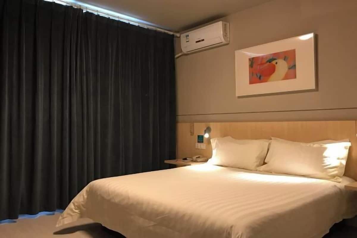 A hotel room with a bed, a nightstand, a wall-mounted air conditioner, and a painting.