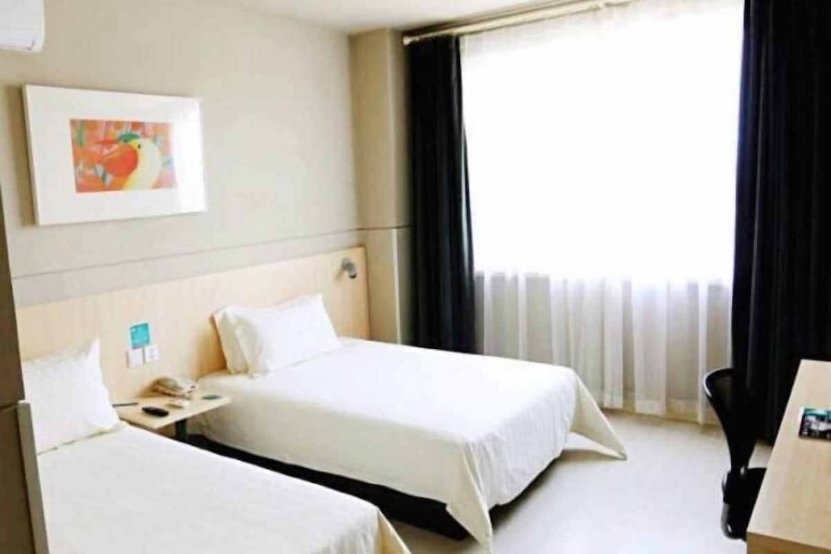 A hotel room with two beds, a desk, a chair, a painting on the wall, and a window with curtains.