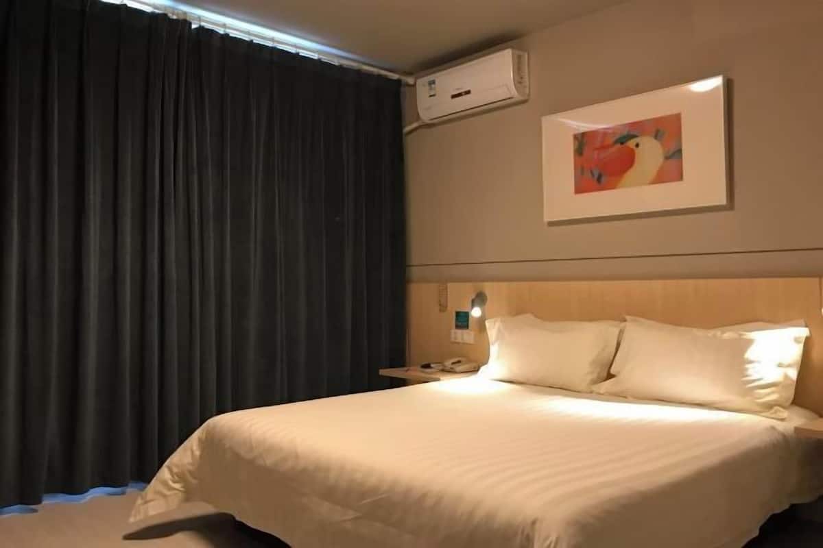 A hotel room with a bed, a nightstand, a wall-mounted air conditioner, and a painting.