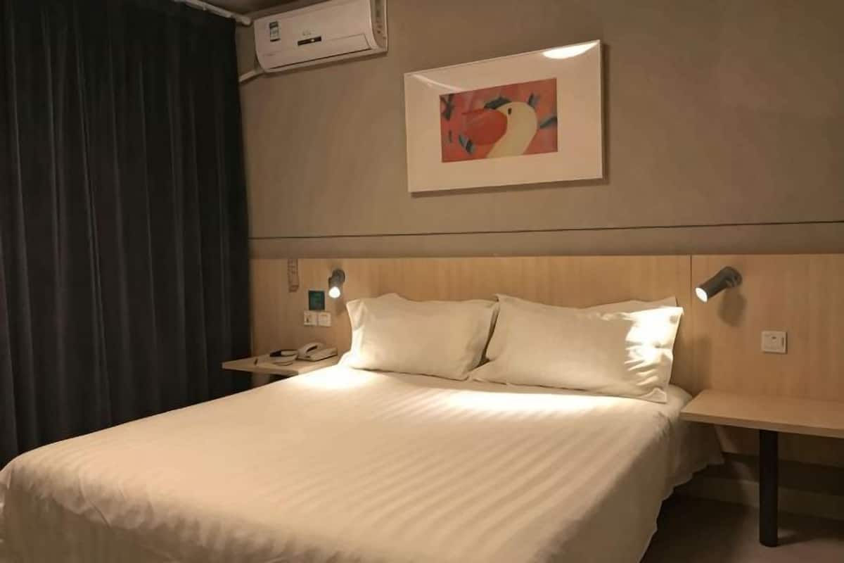 A hotel room with a bed, a nightstand, a wall-mounted air conditioner, and a painting.