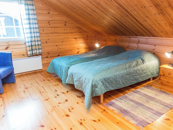 A cozy room with a wooden sloped ceiling, a bed with a quilted cover, a blue chair, and a small bedside table with a lamp.