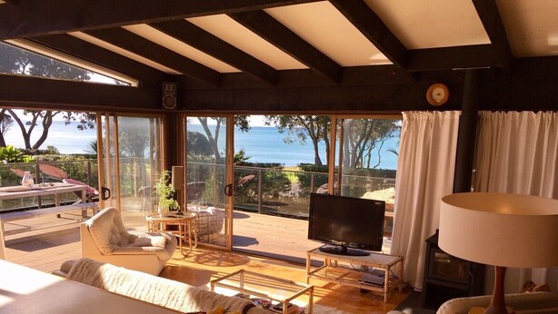 A spacious living room with a large TV, a dining table with fruit, a sofa, and a view of the ocean. - Whiritoa -Glorious Beach Front! (Whiritoa)