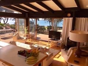 A spacious living room with a large TV, a dining table with fruit, a sofa, and a view of the ocean. - Whiritoa -Glorious Beach Front! (Whiritoa)