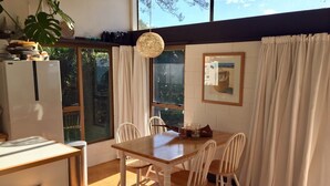 A dining area with a wooden table and chairs, a hanging light fixture, a refrigerator, and a window with curtains. - Whiritoa -Glorious Beach Front! (Whiritoa)
