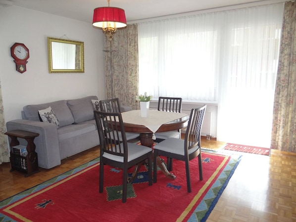 A living room with a sofa, a dining table with chairs, a red lamp, a clock, and a mirror on the wall.