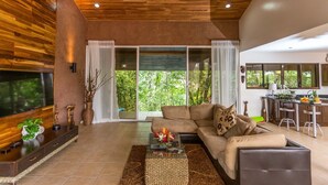 A modern living room with a large TV, a sofa, a coffee table, and a kitchen area with a bar. - Tropical Treehouse with Private Pool - 3BR/2.5B (Manuel Antonio)