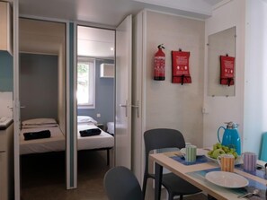Habitación pequeña con cama, una mesa, un plato, tazas y una tetera. Hay dos extintores rojos en la pared.