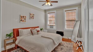 A bedroom with a bed, a ceiling fan, a window with blinds, a small table with a plant, and a whiteboard. - Cozy On Creswell (Shreveport)