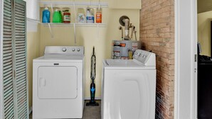 A laundry room with two white washers and dryers, a ceiling fan, and shelves with laundry supplies. - Cozy On Creswell (Shreveport)