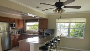 Uma cozinha ampla com uma ilha grande, banquetas, um ventilador de teto e vista para o verde lá fora.