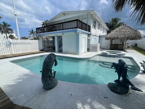 Une piscine ornée de deux statues de dauphins, une maison blanche dotée d’un balcon et une construction à toit de chaume.