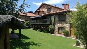 A two-story stone house with a thatched-roof structure, a balcony with potted plants, and a well-maintained lawn.