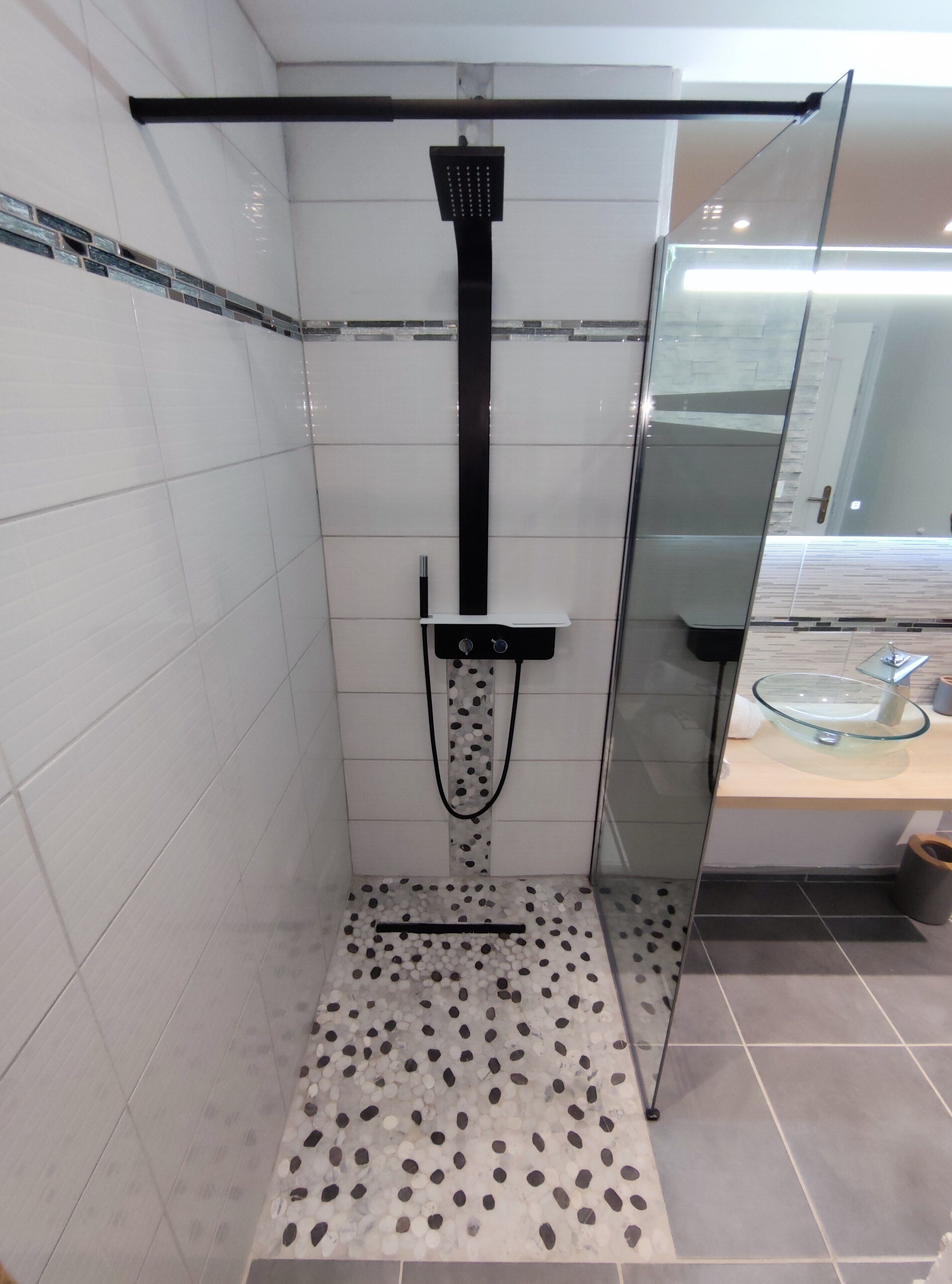 A modern bathroom with a glass shower enclosure, white tiled walls, and a pebble-patterned floor.