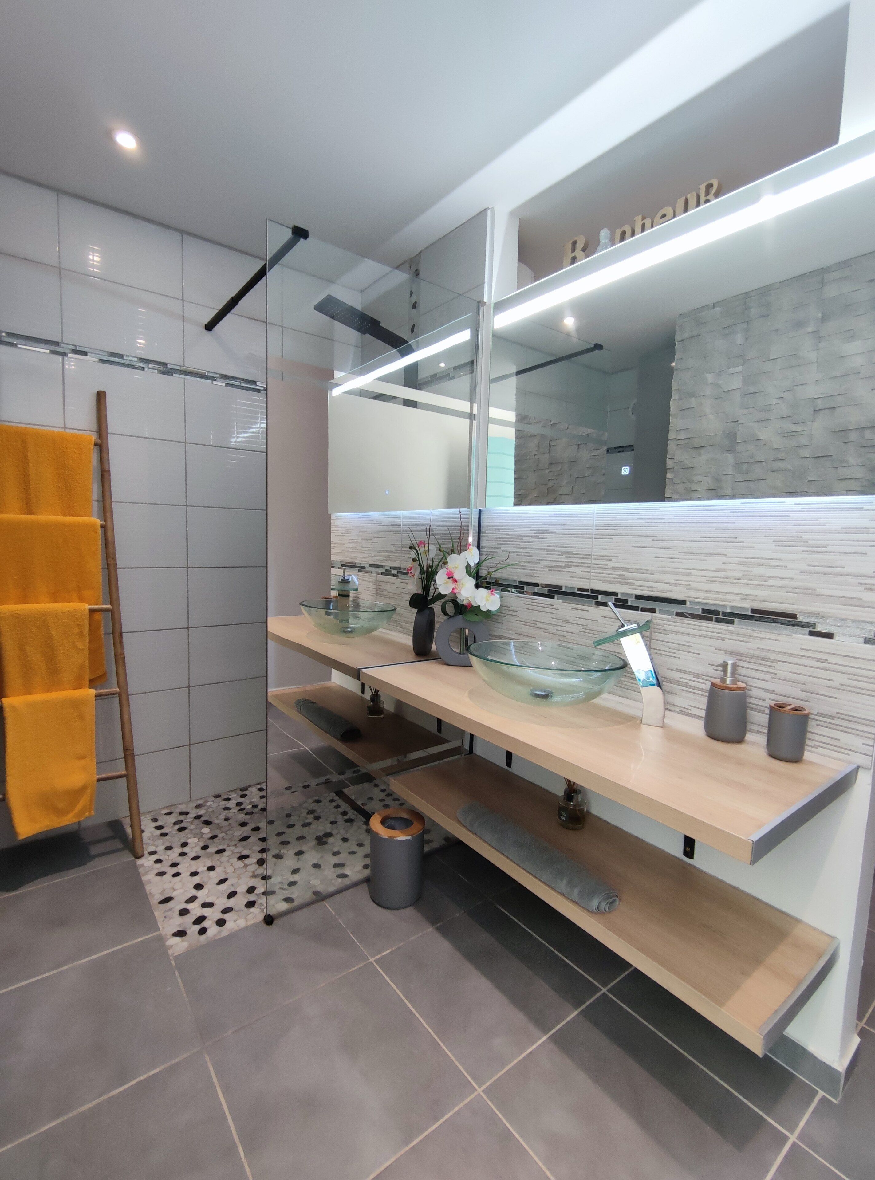 Un baño moderno con lavamanos de vidrio, estantes de madera, un espejo grande y un perchero con toallas amarillas.