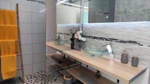 A modern bathroom with a glass sink, wooden shelves, a large mirror, and a towel rack with yellow towels. - Gray Diamond Studio (Le Diamant)