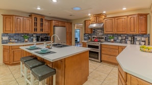 A modern kitchen with wooden cabinets, a central island with bar stools, stainless steel appliances, and a backsplash with blue tiles. - ADA Access, One Story, Htd POOL, Spa, Putting Green, Games, Fire Pit, TVs All BRs (Scottsdale)