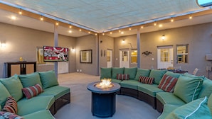 A spacious outdoor living area with a curved green sofa set, a central fire pit, and a television mounted on the wall. - ADA Access, One Story, Htd POOL, Spa, Putting Green, Games, Fire Pit, TVs All BRs (Scottsdale)