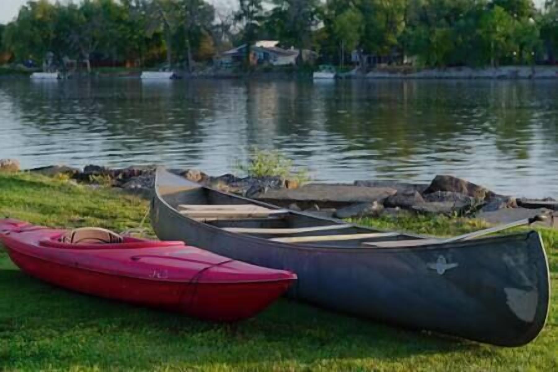 Una fila de canoas y un kayak junto a un lago tranquilo con orillas cubiertas de césped y árboles al fondo.