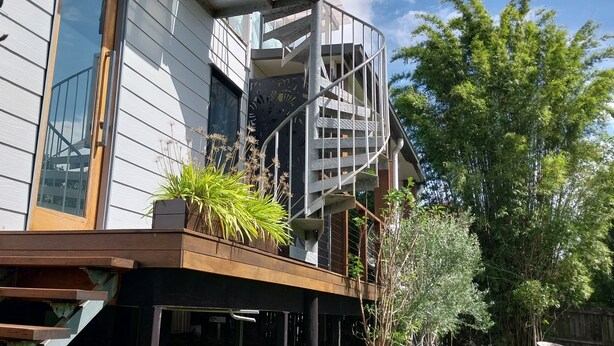 A modern building with a wooden deck, a spiral staircase, and a large tree in the background. - The Sky House by the bay (Wynnum)