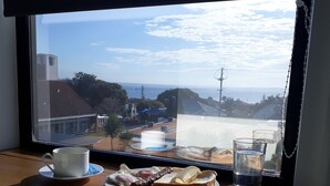 A breakfast tray with eggs, sausage, and toast on a wooden table by a window overlooking a coastal view. - The Sky House by the bay (Wynnum)
