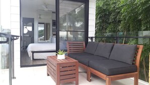 A balcony with a wooden sofa, a small table with a plant, and a bed visible through a glass door. - The Sky House by the bay (Wynnum)