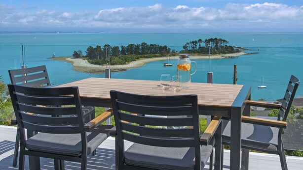 Una mesa y sillas de madera en una terraza con vista al mar turquesa y una pequeña isla.