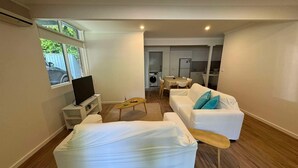 A modern living room with a sofa, a coffee table, and a TV. There is a dining area with a table and chairs. The room has wooden flooring and white walls. - 3 Bedroom, pet friendly unit, 2min walk from main street, beach and supermarket. (Lorne)