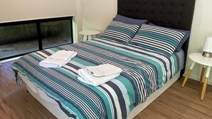 A neatly made bed with striped bedding and two pillows, a small bedside table with a lamp, and a window with blinds. - 3 Bedroom, pet friendly unit, 2min walk from main street, beach and supermarket. (Lorne)