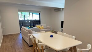 A modern living room with a white sofa set, a dining table with white chairs, and a large window with a view of greenery. - 3 Bedroom, pet friendly unit, 2min walk from main street, beach and supermarket. (Lorne)