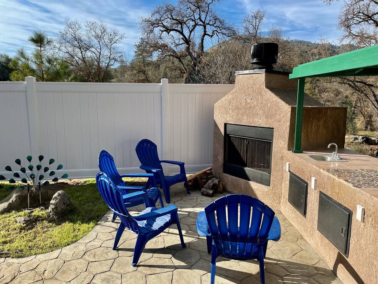 Une terrasse extérieure avec des chaises bleues, un sol carrelé en pierre, une clôture blanche et une cheminée en pierre.