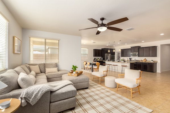 A modern living room with a sectional sofa, a dining area, and a kitchen with dark cabinetry and stainless steel appliances.