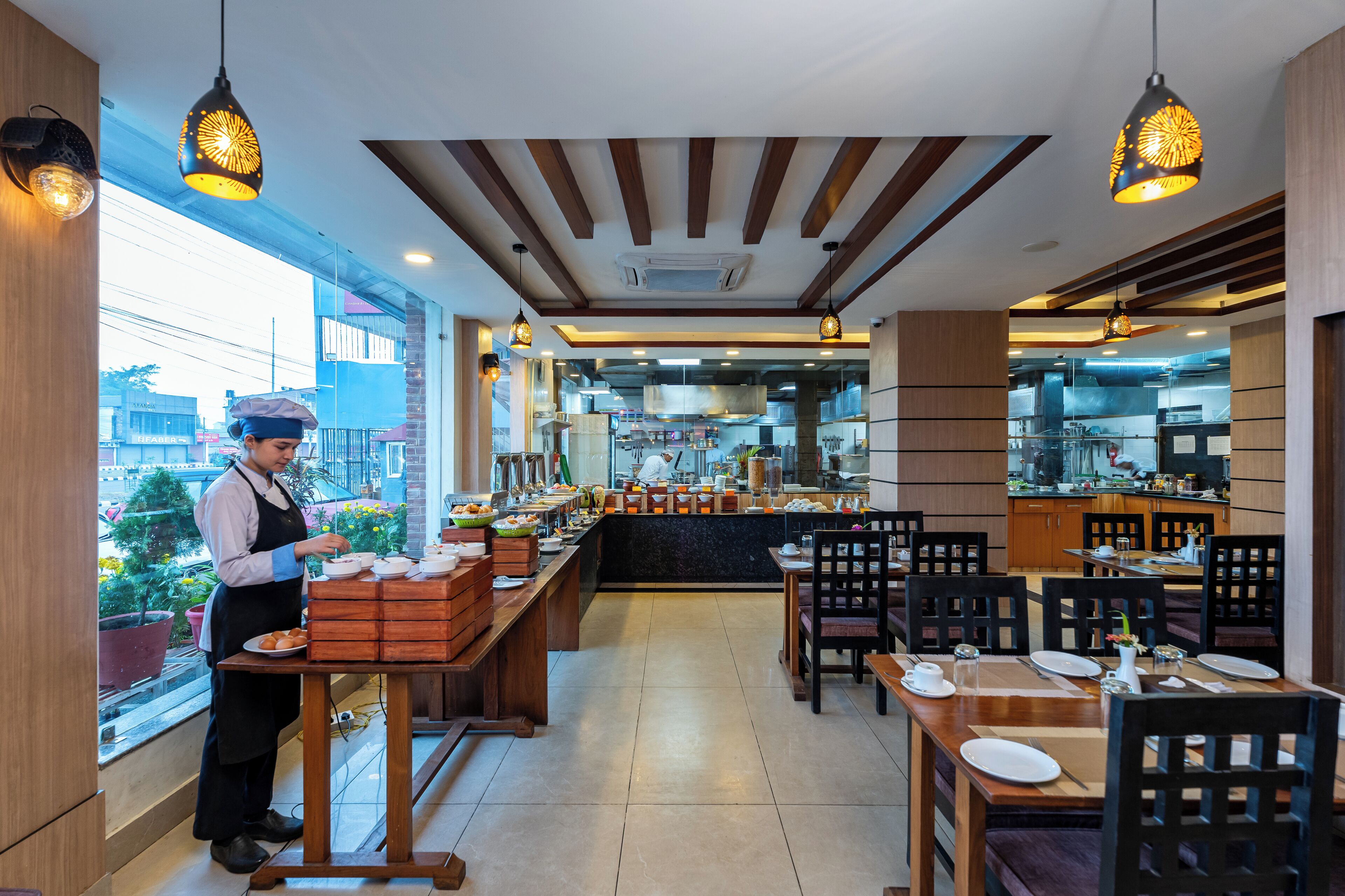 A restaurant with a chef preparing food, wooden tables and chairs, and a view of the kitchen through the glass wall.