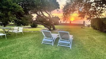 A grassy outdoor area with two lounge chairs and a table, surrounded by trees and a sunset view.