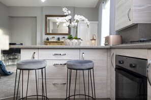 A modern kitchen with two bar stools, a large island with a vase of white flowers, a stove, and a microwave. - Stylish George Street Apartment (Peebles)