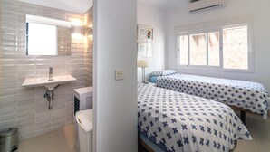 Et værelse med håndvask, to senge med mønstret sengetøj, et vindue og en aircondition-enhed.