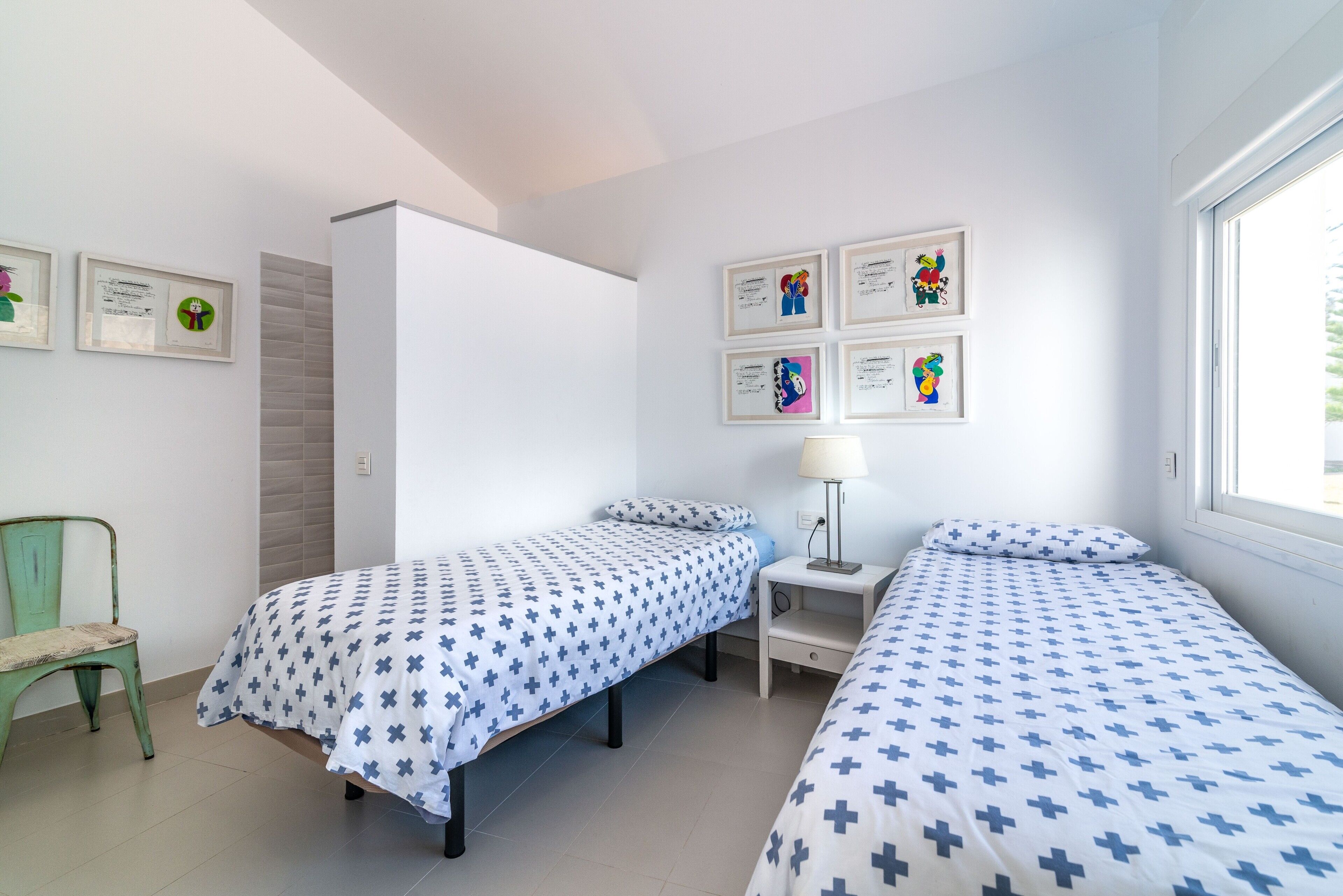 A room with two beds, a chair, a nightstand with a lamp, and framed artwork on the wall.
