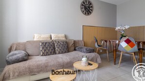 Un salon comprenant un canapé, une table basse ronde en bois, une horloge et un coin repas avec des chaises et une table.