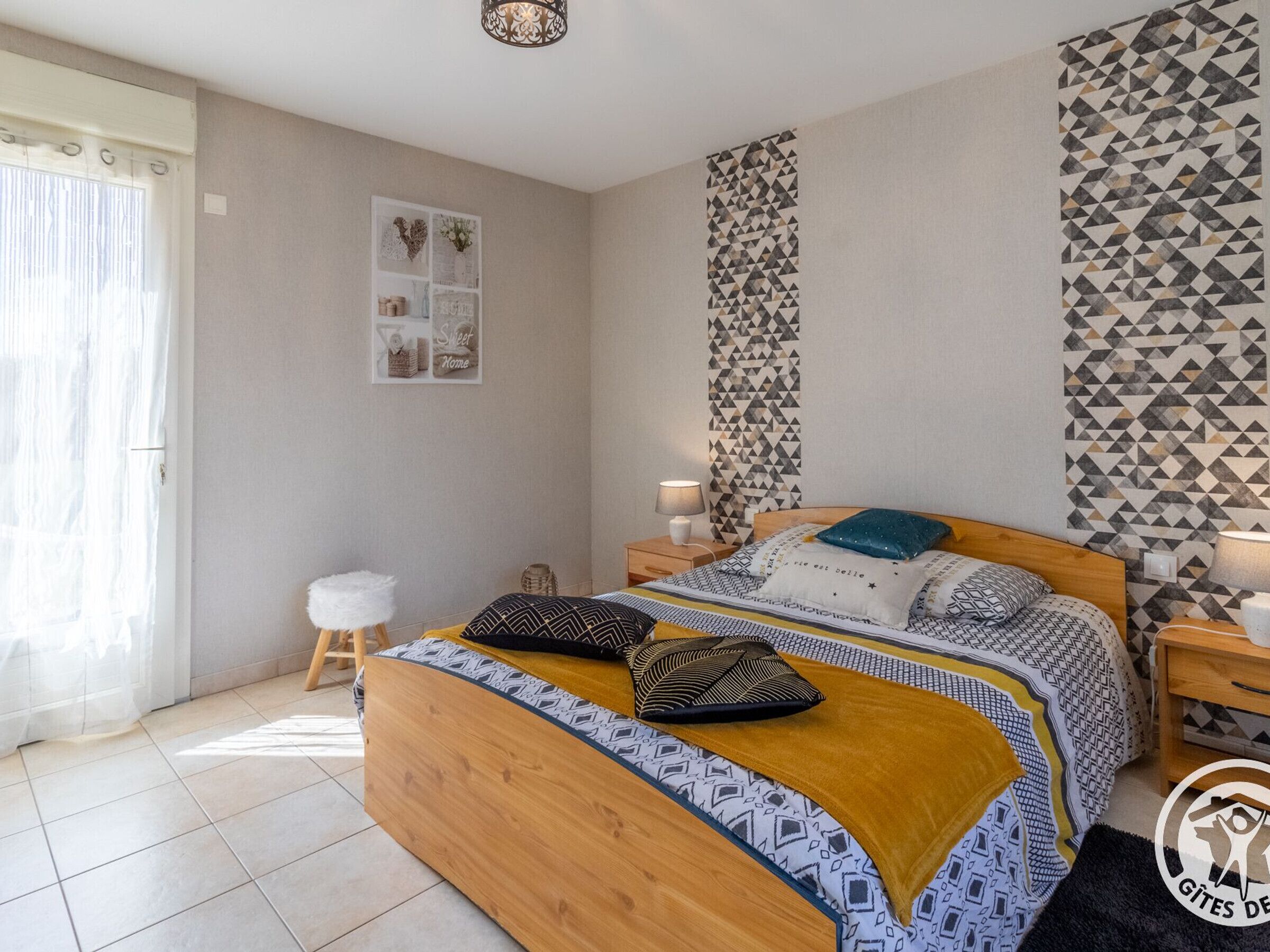 Een slaapkamer met een houten bed, beddengoed met een patroon, een nachtkastje, een muur met geometrisch behang en een klein krukje.