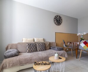 Una sala de estar con un sofá, una mesa redonda de madera, un reloj y una zona de comedor con sillas y una mesa.