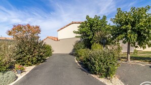 Eine Auffahrt führt zu einem Gebäude mit einem Satteldach und einer Garage. Bäume und Sträucher säumen die beiden Seiten der Auffahrt.