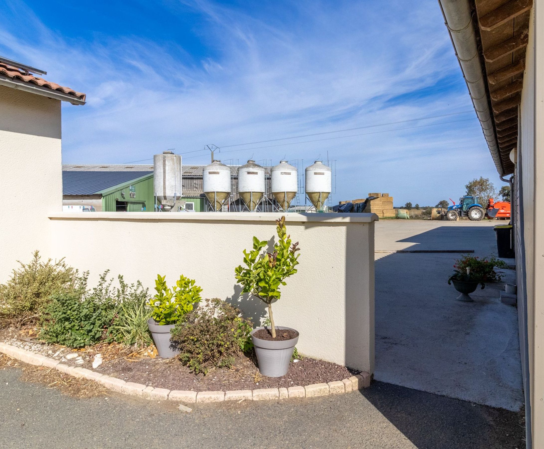 Un bâtiment à l’extérieur blanc, un petit jardin agrémenté de plantes en pot et, au loin, une vue sur du matériel agricole et des silos.