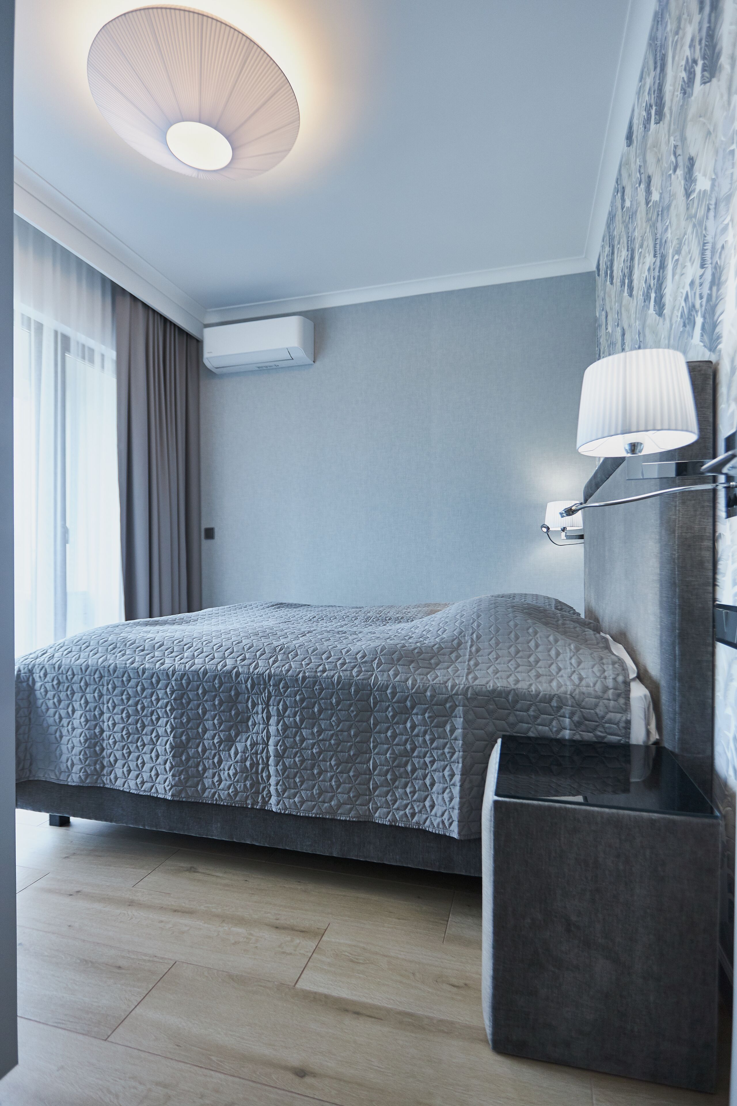 A modern hotel room with a bed, bedside table, lamp, air conditioning unit, and a patterned wall.