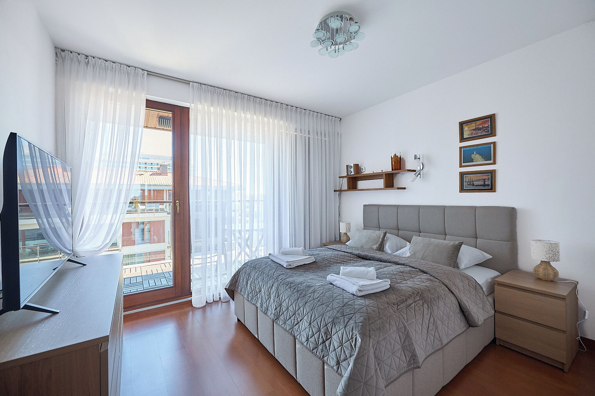A bedroom with a bed, bedside table, curtains, a TV, and a balcony.
