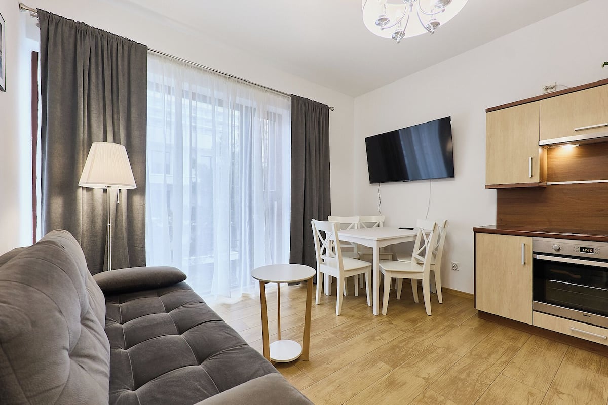 Ett modernt vardagsrum med en grå soffa, ett litet runt bord och en matplats med vita stolar. En platt-TV är monterad på väggen, och det finns ett köksområde med träskåp och en ugn.